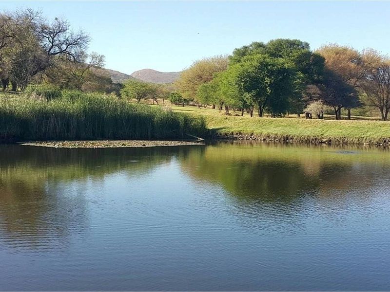A scenic view of a natural pond surrounded by lush greenery, mature trees, and rolling hills in the background.