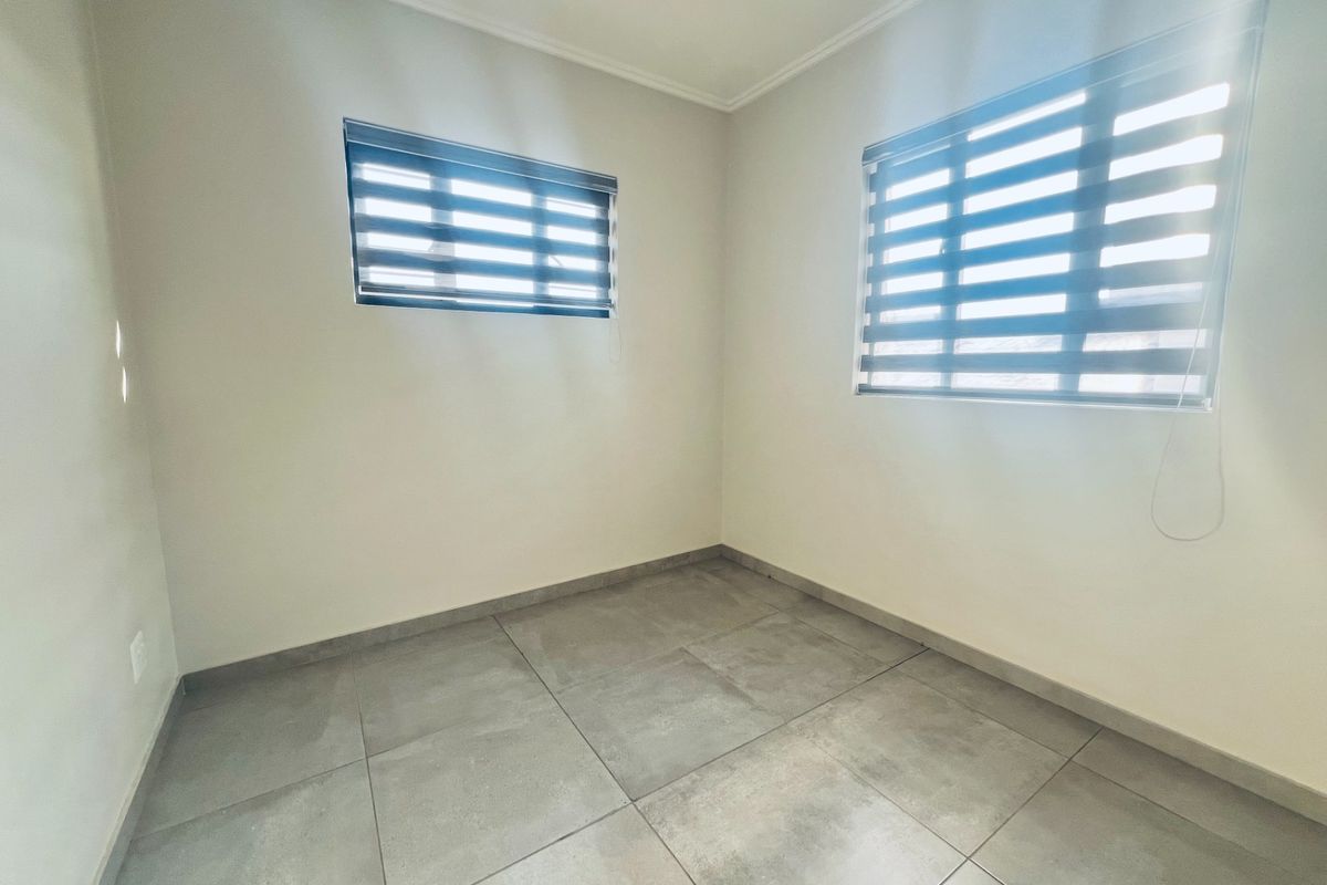 A bright, empty bedroom featuring neutral walls, large format grey floor tiles, and modern zebra blinds on the windows.