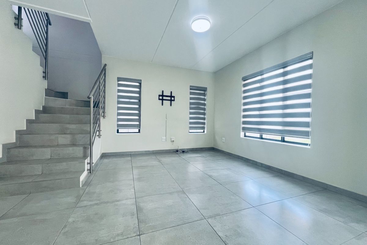 A bright, open-plan living area featuring modern grey floor tiles, a staircase with stainless steel railings, and zebra blinds on the windows.