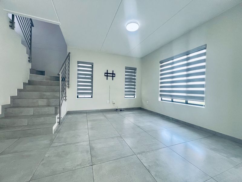 A bright, open-plan living area featuring modern grey floor tiles, a staircase with stainless steel railings, and zebra blinds on the windows.