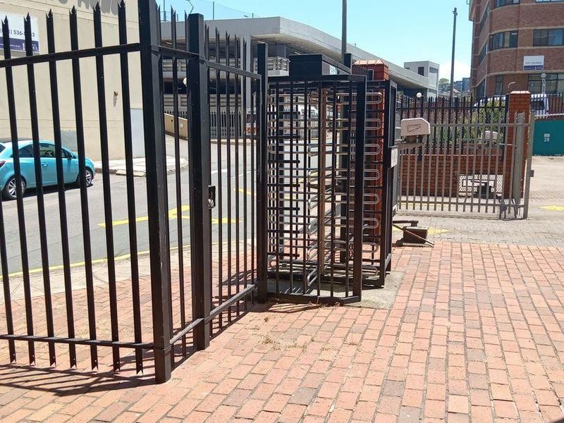 Commercial property entrance featuring a secure pedestrian turnstile, steel perimeter fencing, and brick paved access area.