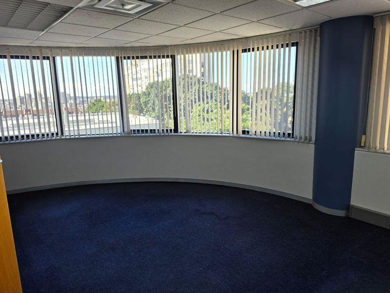 An empty, curved office space featuring large windows with vertical blinds, a suspended ceiling, and blue carpeting, offering an open layout for professional use.