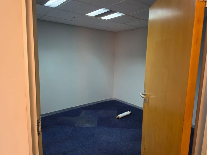 An empty commercial office space featuring blue carpet tiles, a suspended ceiling with recessed lighting, and a solid wood door.