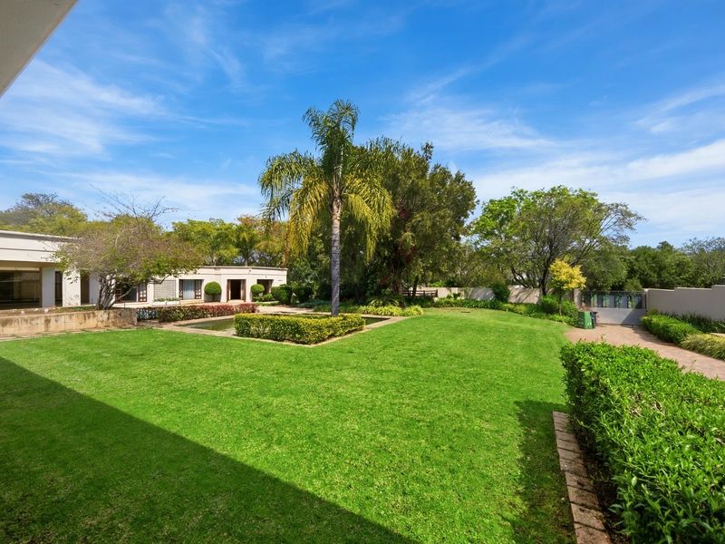 A spacious, well-maintained rear garden featuring a manicured lawn, mature palm tree, water feature, and paved driveway access.