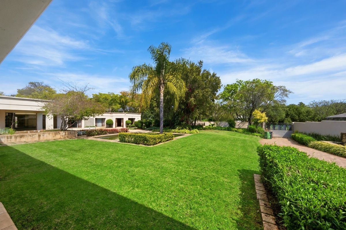 A spacious, well-maintained rear garden featuring a manicured lawn, mature palm tree, water feature, and paved driveway access.