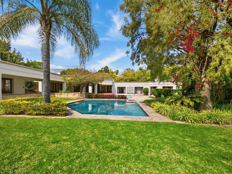 A well-maintained outdoor pool area featuring a paved patio, lush garden landscaping, and a covered veranda attached to the main residence.