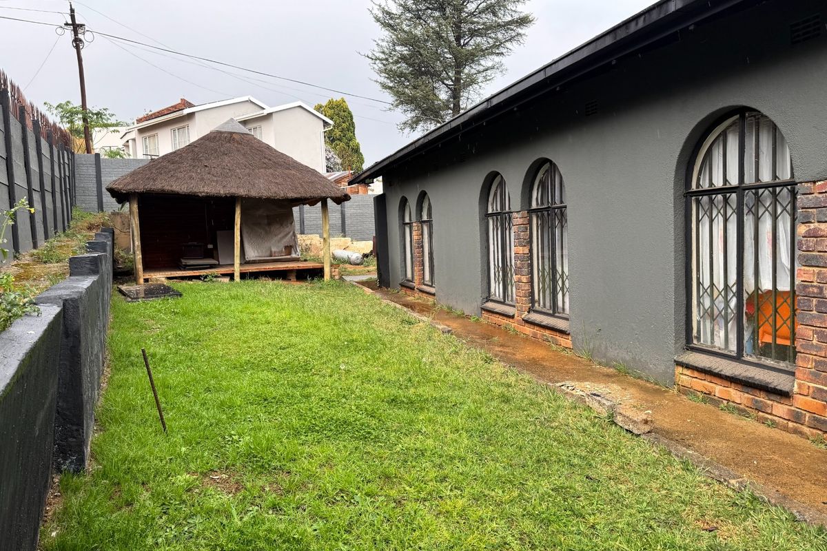 Rear garden area featuring a thatched lapa, concrete pathway, and arched windows along the side of the house with security bars.