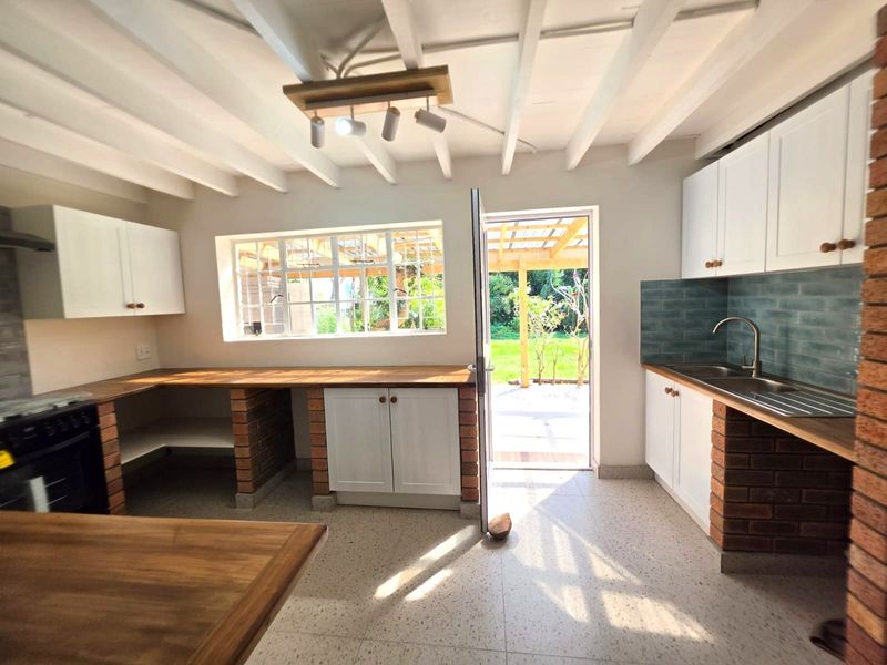 A bright, rustic-style kitchen featuring exposed white beams, brick-based cabinetry with wooden countertops, and direct access to the rear garden.