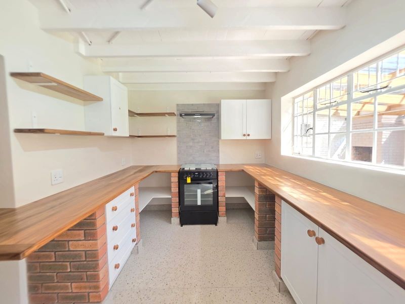 A bright, modern kitchen featuring exposed timber ceiling beams, brick-based cabinetry with wooden countertops, a freestanding stove, and durable terrazzo flooring.