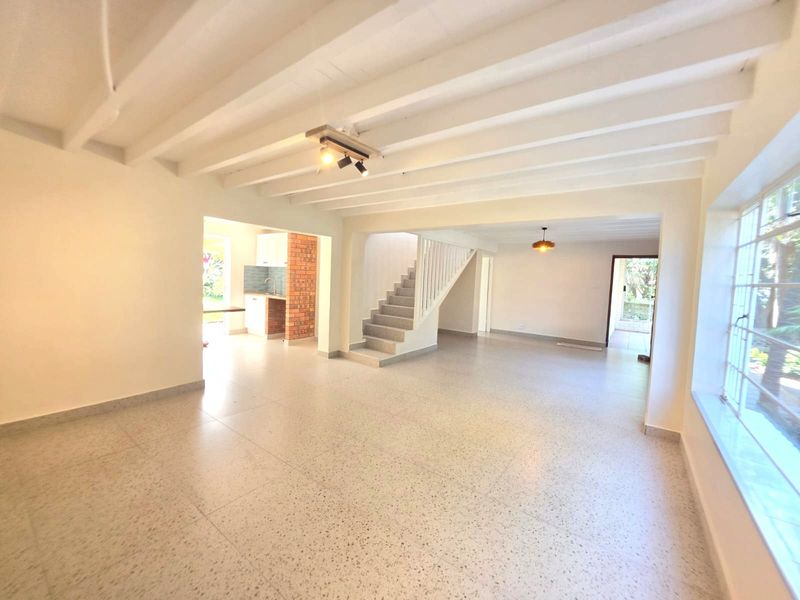 Spacious open-plan living area featuring exposed beam ceilings, terrazzo flooring, and a staircase, with a glimpse into the kitchen area.