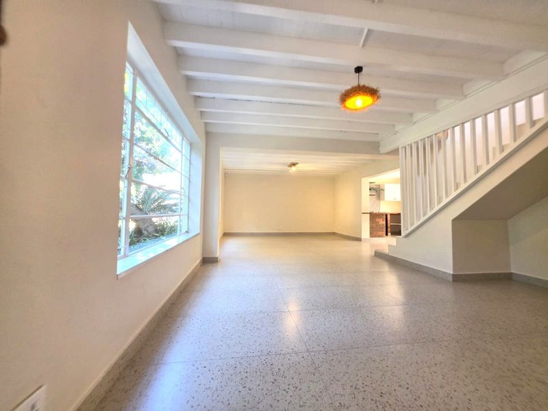 Spacious open-plan living area featuring exposed white timber beams, classic terrazzo flooring, and a staircase, offering a bright and versatile layout.