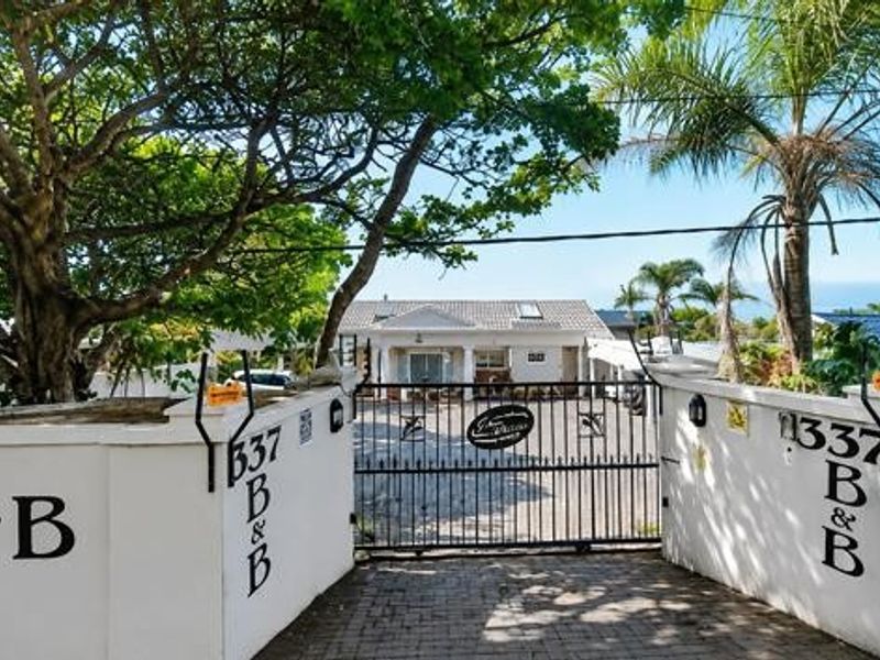 Front entrance view of the property featuring a secure wrought iron gate, masonry boundary walls, and a paved driveway leading to the main house.