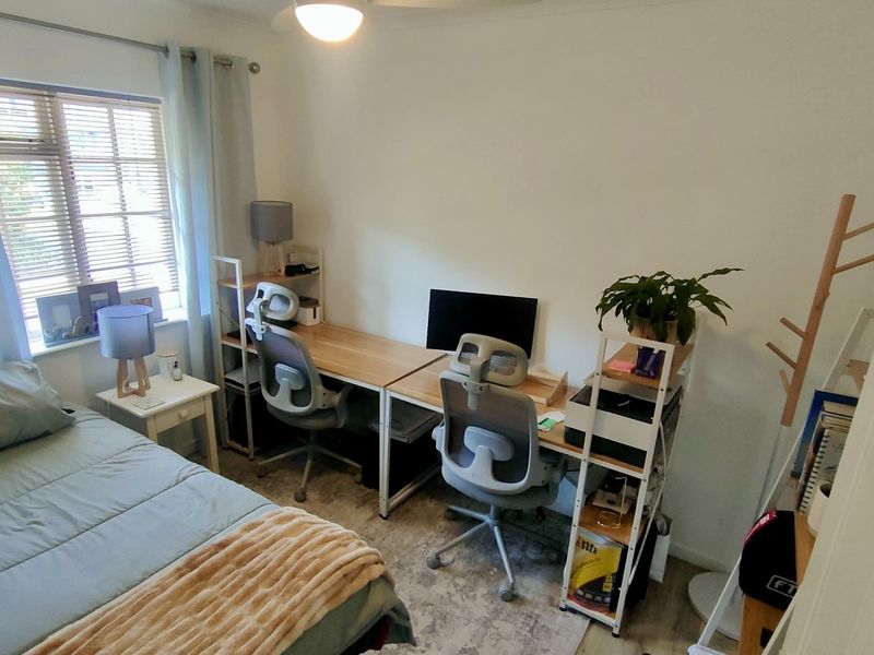 A functional bedroom space featuring a ceiling fan and window blinds, currently utilized as a combined sleeping and home office area.