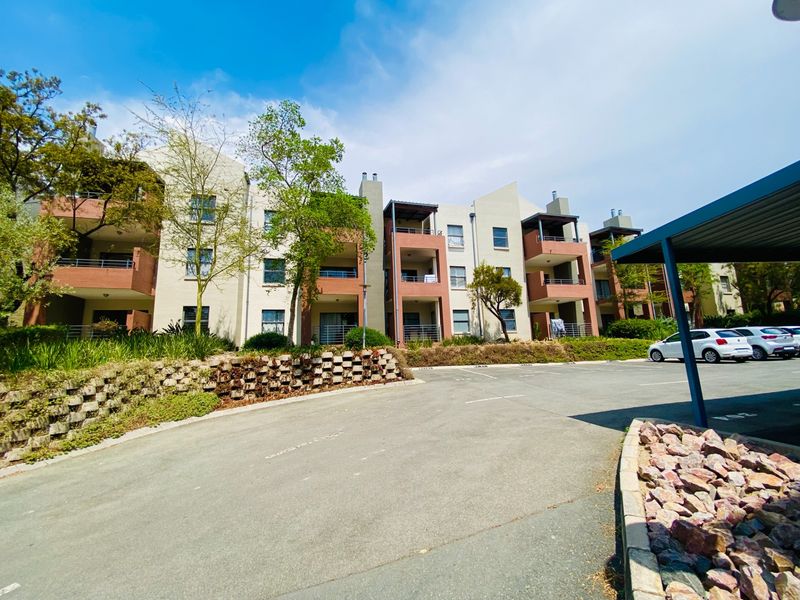 Exterior view of a multi-story apartment complex featuring balconies, landscaped garden areas, and a paved parking lot with covered bays.