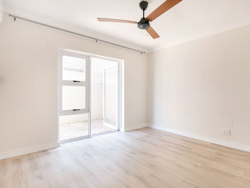 A bright, empty bedroom featuring light laminate flooring, a ceiling fan, and direct access to an outdoor area through a glass door.