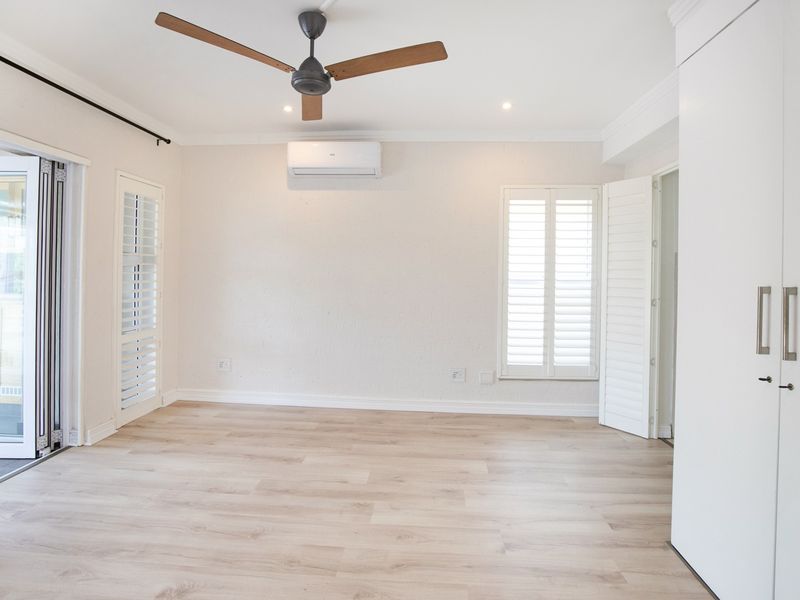 A bright, empty bedroom featuring light laminate flooring, built-in cupboards, plantation shutters, a ceiling fan, and a split-system air conditioner.