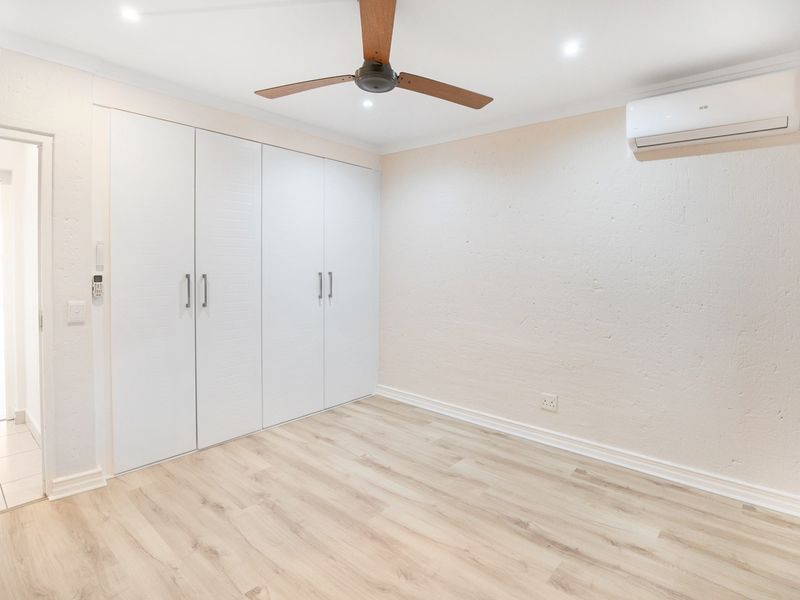 A bright, empty bedroom featuring modern laminate flooring, built-in cupboards, a ceiling fan, and a wall-mounted split-system air conditioner.