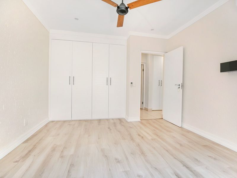 A bright, empty bedroom featuring modern laminate flooring, built-in cupboards, a ceiling fan, and an integrated air conditioning unit.