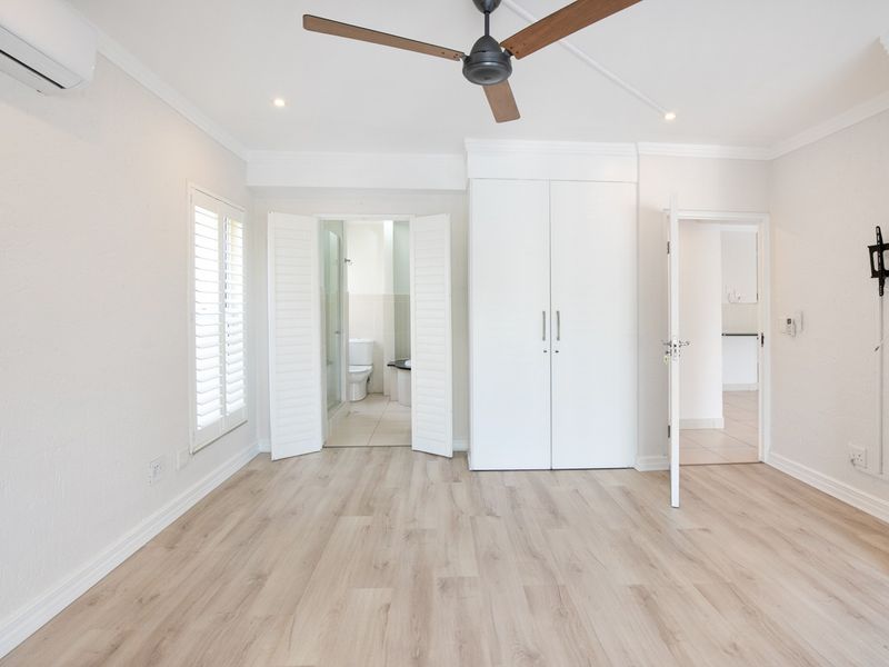 A bright, empty bedroom featuring light laminate flooring, built-in cabinetry, a ceiling fan, and an integrated air conditioning unit with access to an en-suite bathroom.