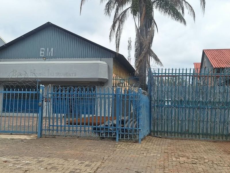 Front exterior view of a commercial industrial property featuring a corrugated metal structure, secure perimeter fencing with razor wire, and a paved entrance area.