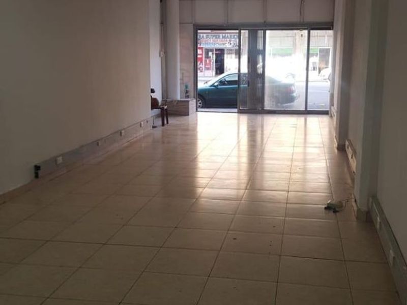 An empty commercial open-plan space featuring tiled flooring, a suspended ceiling with recessed lighting, and a large sliding glass entrance door.