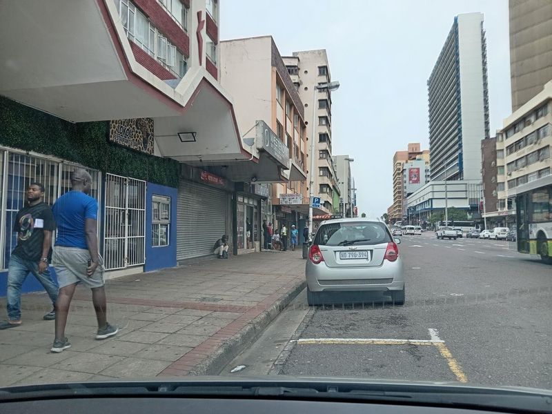 A street-level view of a commercial building facade featuring storefronts with security roller shutters along a paved city sidewalk.