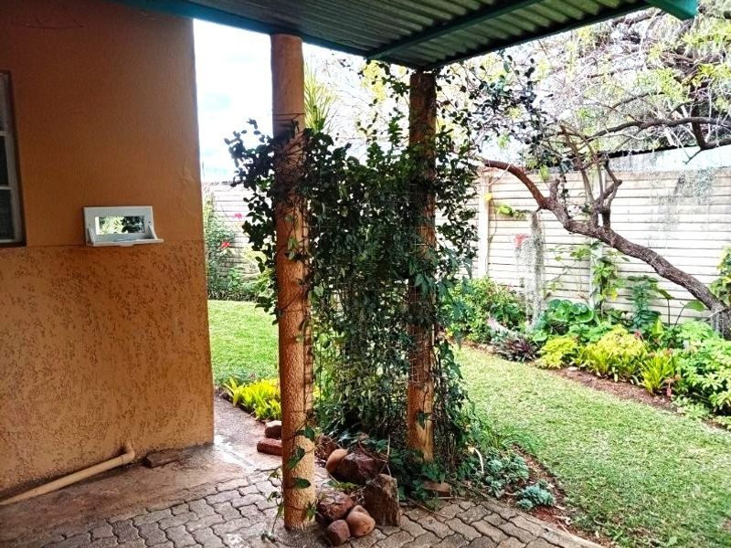 A covered patio area featuring paved flooring and structural columns, overlooking a private garden with established landscaping and boundary walling.