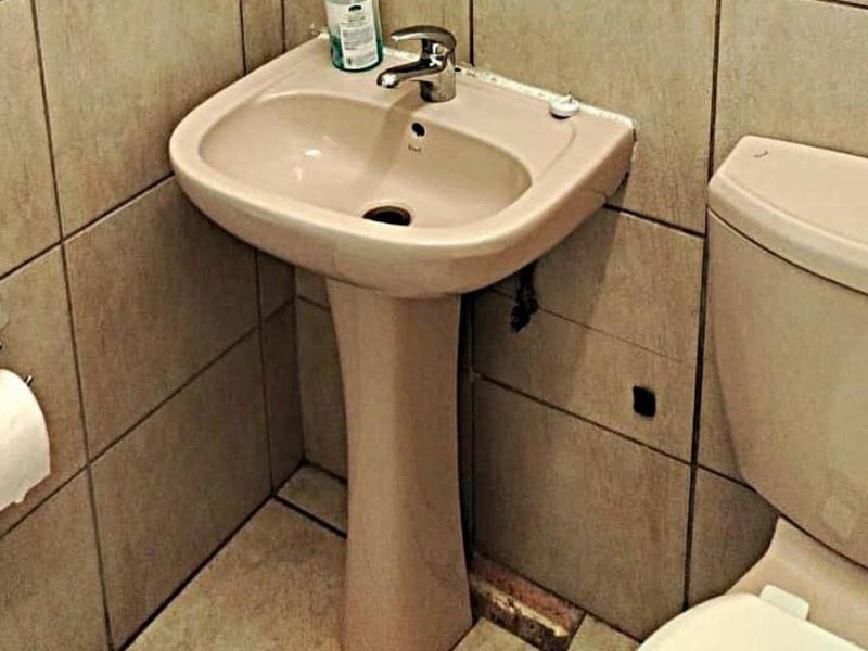 A compact bathroom featuring a pedestal basin, ceramic toilet, and neutral wall tiling. The space requires minor repairs to floor tiles and plumbing fixtures.