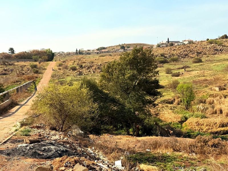 A view of the vacant residential land featuring a dirt access road, natural vegetation, and rocky terrain with surrounding neighborhood in the distance.