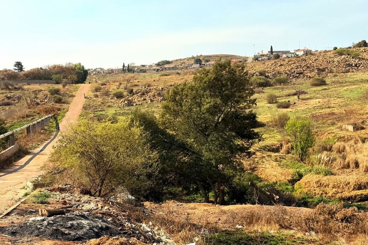 A view of the vacant residential land featuring a dirt access road, natural vegetation, and rocky terrain with surrounding neighborhood in the distance.