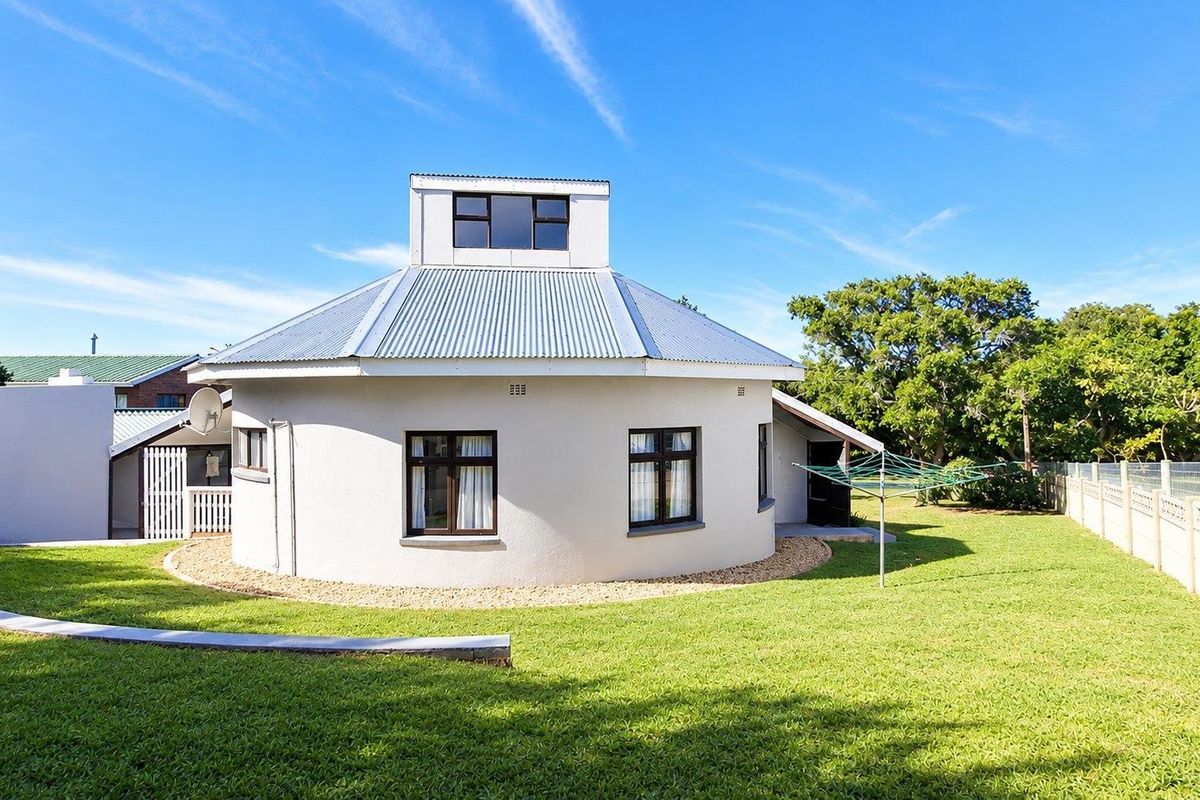 Rear view of a unique circular house featuring a corrugated metal roof, clerestory windows, and a well-maintained lawn with boundary walling.