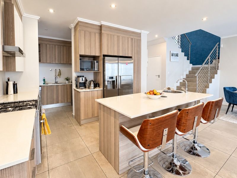 Modern open-plan kitchen featuring wood-finish cabinetry, stone countertops, a central island with sink, and integrated appliances, flowing into a dining area with staircase access.