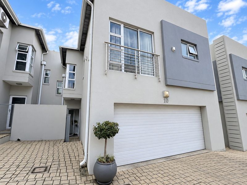 Modern double-story townhouse facade featuring a paved driveway, automated garage door, and an upper-level balcony with stainless steel railing.