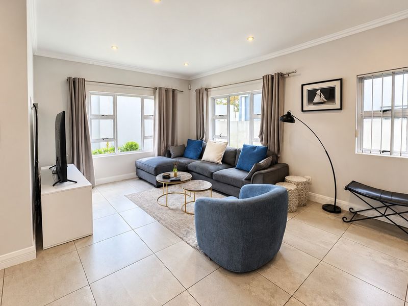 A bright and well-maintained living room featuring neutral tiled flooring, recessed lighting, and large aluminium-framed windows with security bars.