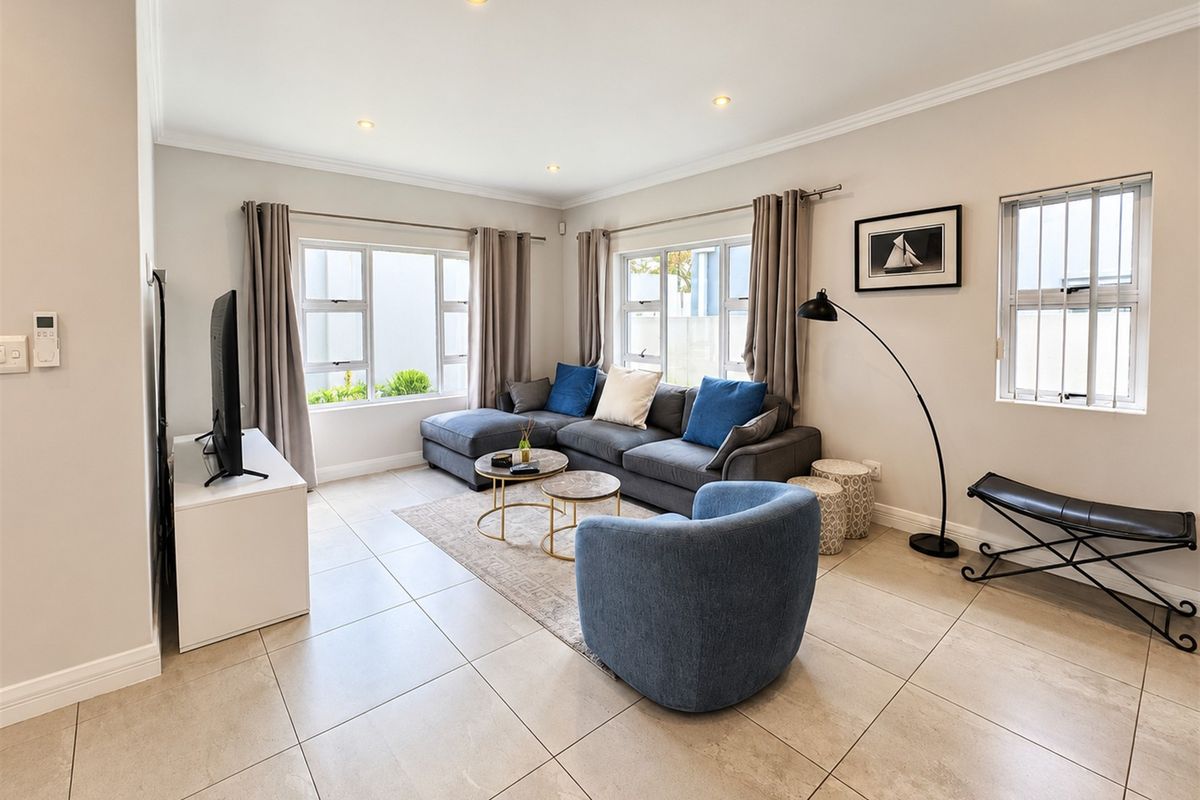 A bright and well-maintained living room featuring neutral tiled flooring, recessed lighting, and large aluminium-framed windows with security bars.