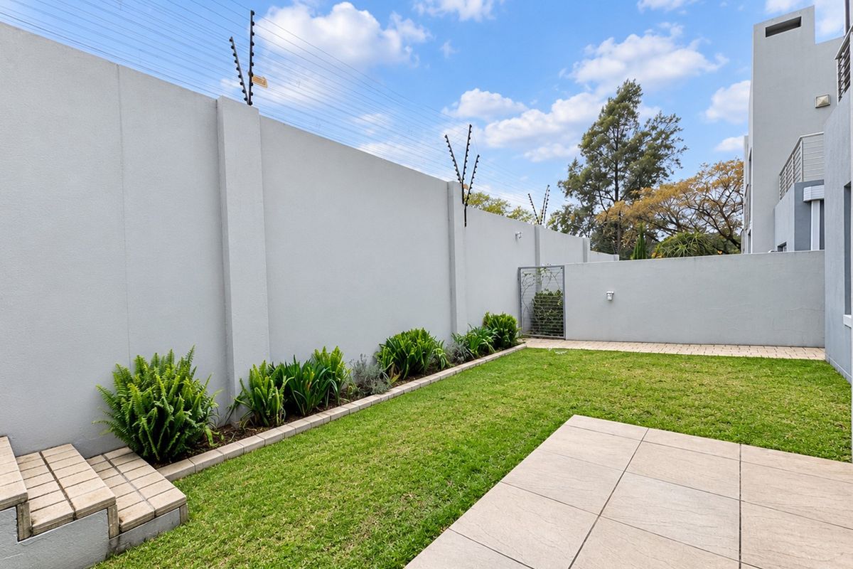 A well-maintained rear garden featuring a paved patio area, manicured lawn, landscaped garden beds, and secure perimeter walling with electric fencing.