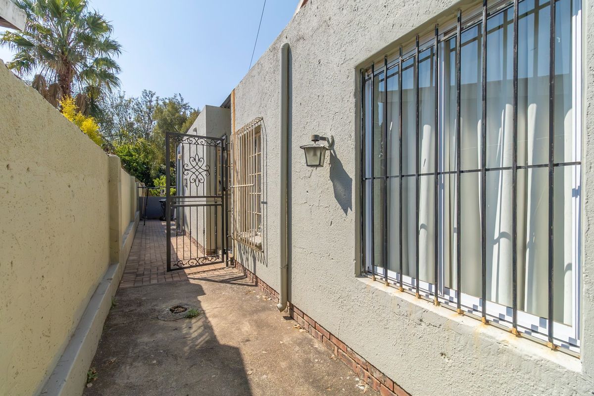A narrow rear exterior walkway featuring paved flooring, security gates, and protective window bars along the side of the property.