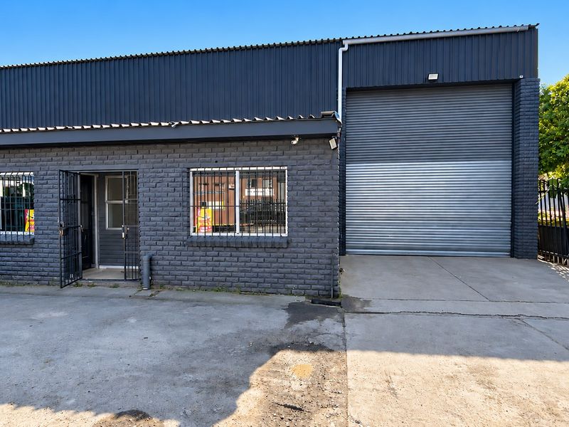 Industrial property featuring a dark brick facade, large roller shutter door, security bars on windows, and a concrete driveway.