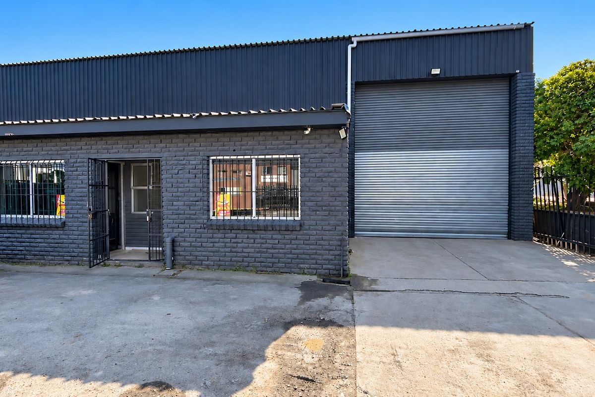 Industrial property featuring a dark brick facade, large roller shutter door, security bars on windows, and a concrete driveway.