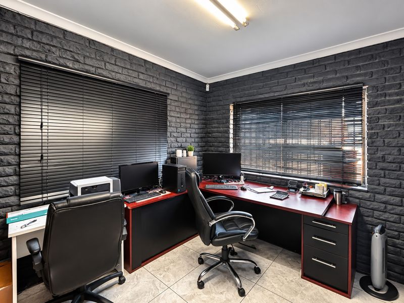 A well-maintained home office featuring dark exposed brick walls, tiled flooring, and large windows fitted with venetian blinds.