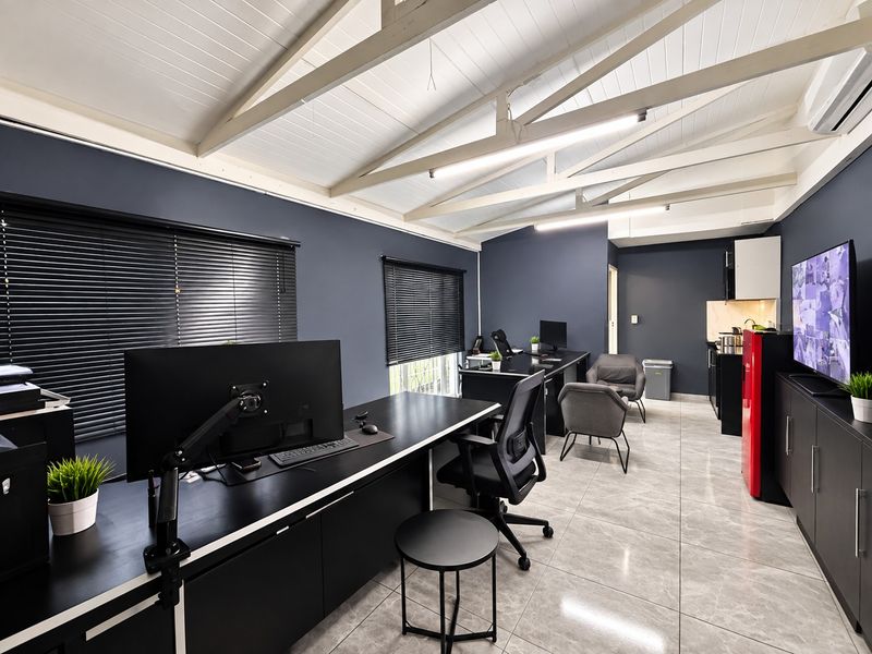 Modern industrial-style office space featuring exposed white timber trusses, sleek tiled flooring, dark feature walls, and a compact integrated kitchenette area.