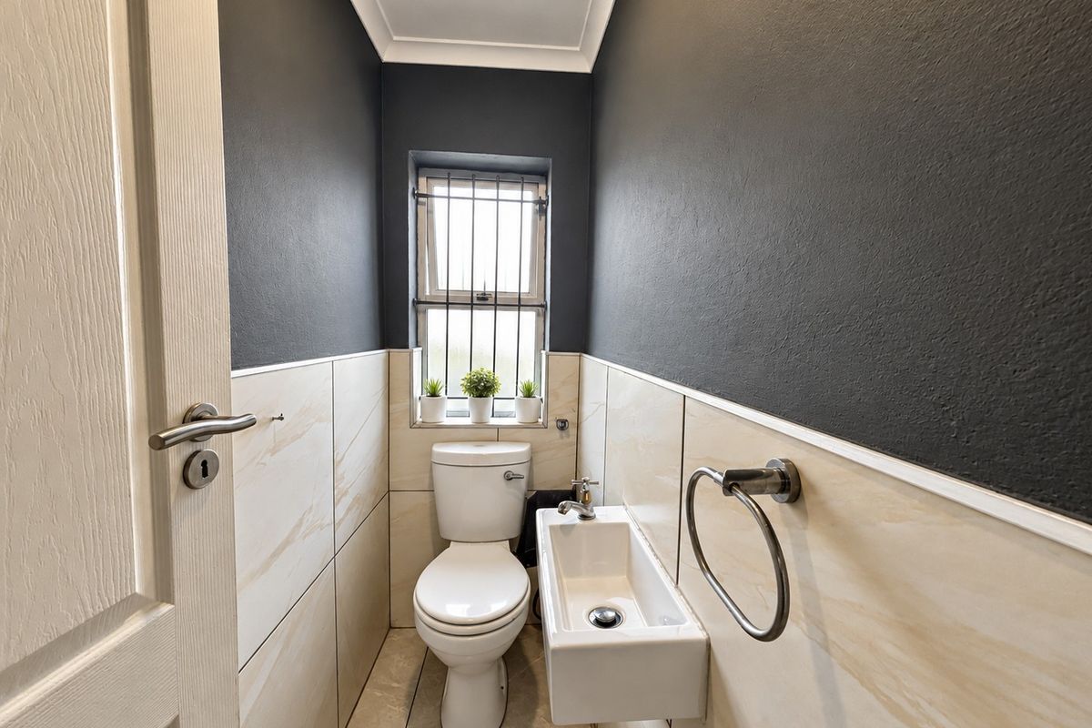 A compact, modern guest bathroom featuring tiled walls, a wall-mounted basin, and a toilet, finished with dark accent walls and security bars on the window.