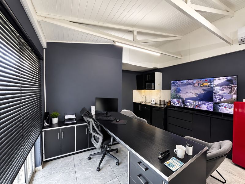 Modern industrial-style office space featuring exposed white timber trusses, dark cabinetry, a compact kitchenette, and tiled flooring.