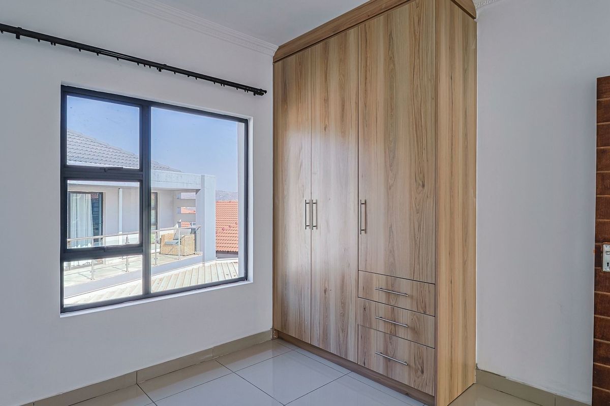 A bright bedroom featuring modern tiled flooring, a large built-in wooden wardrobe, and an aluminium-framed window with views of the surrounding area.