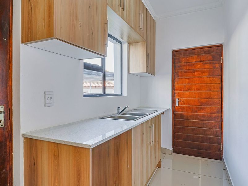 A clean and functional scullery featuring modern wood-finish cabinetry, stone countertops, a stainless steel sink, and tiled flooring.