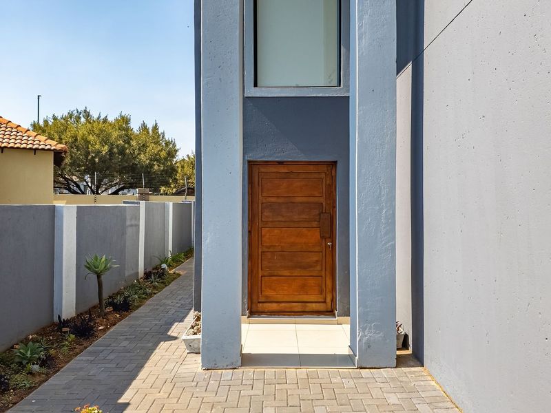 A modern front entrance featuring a solid wooden door, tiled porch, and a paved walkway alongside a clean boundary wall.