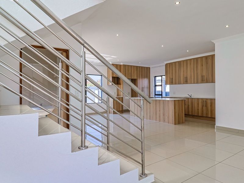 Modern open-plan living area featuring a sleek stainless steel staircase, tiled flooring, and a contemporary kitchen with wood-finish cabinetry and a central island.