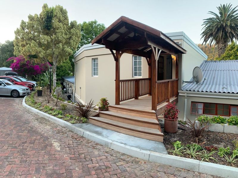 Front exterior view of the property featuring a raised timber entrance deck with a pergola, brick paved driveway, and landscaped garden beds.