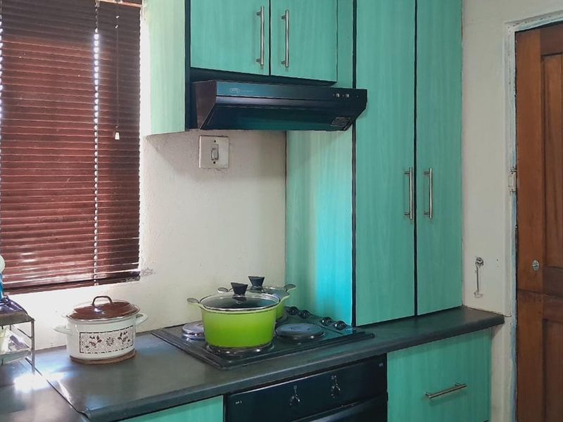 A compact kitchen featuring teal cabinetry, a built-in hob, oven, and extractor fan, with a wooden door and window blinds.