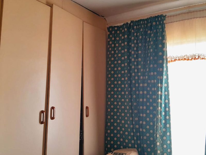 A bedroom featuring built-in cupboards and a window with curtains, showing signs of wear and requiring cosmetic updates.